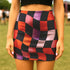 Honey Dazed mini pencil skirt featuring a bold wavy checkerboard pattern in black, orange, purple, and pink. Made from soft eco-polyester with a curve-hugging stretch fit and a flattering high-waist silhouette. The print has a slightly glossy finish, giving it a sleek and modern look. Designed for festival fits, streetwear moments, and retro rave energy. A vivid, sculptural mini skirt that pairs easily with crop tops and confident energy.